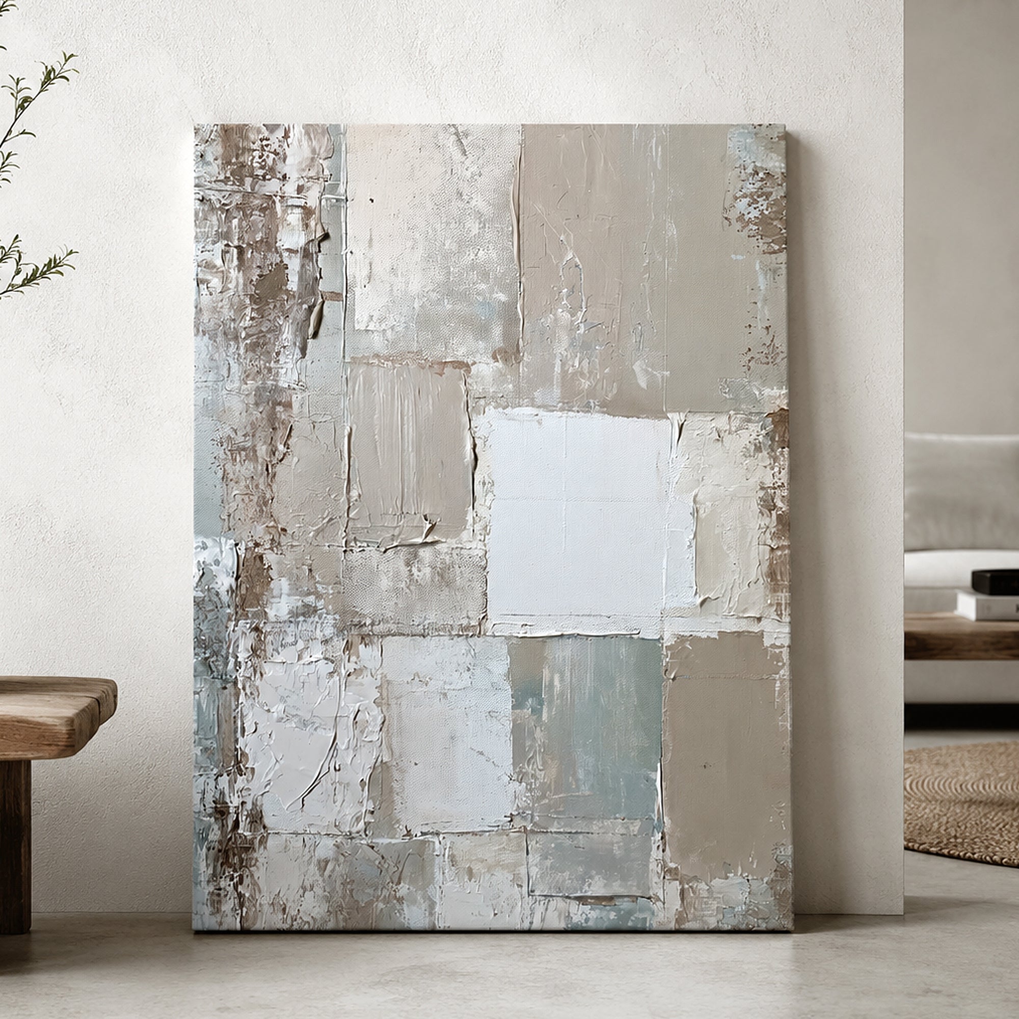 An abstract canvas wall art print with textured brushstrokes in beige, gray, white, and muted blue, leaning against a textured off-white wall in a modern interior setting.