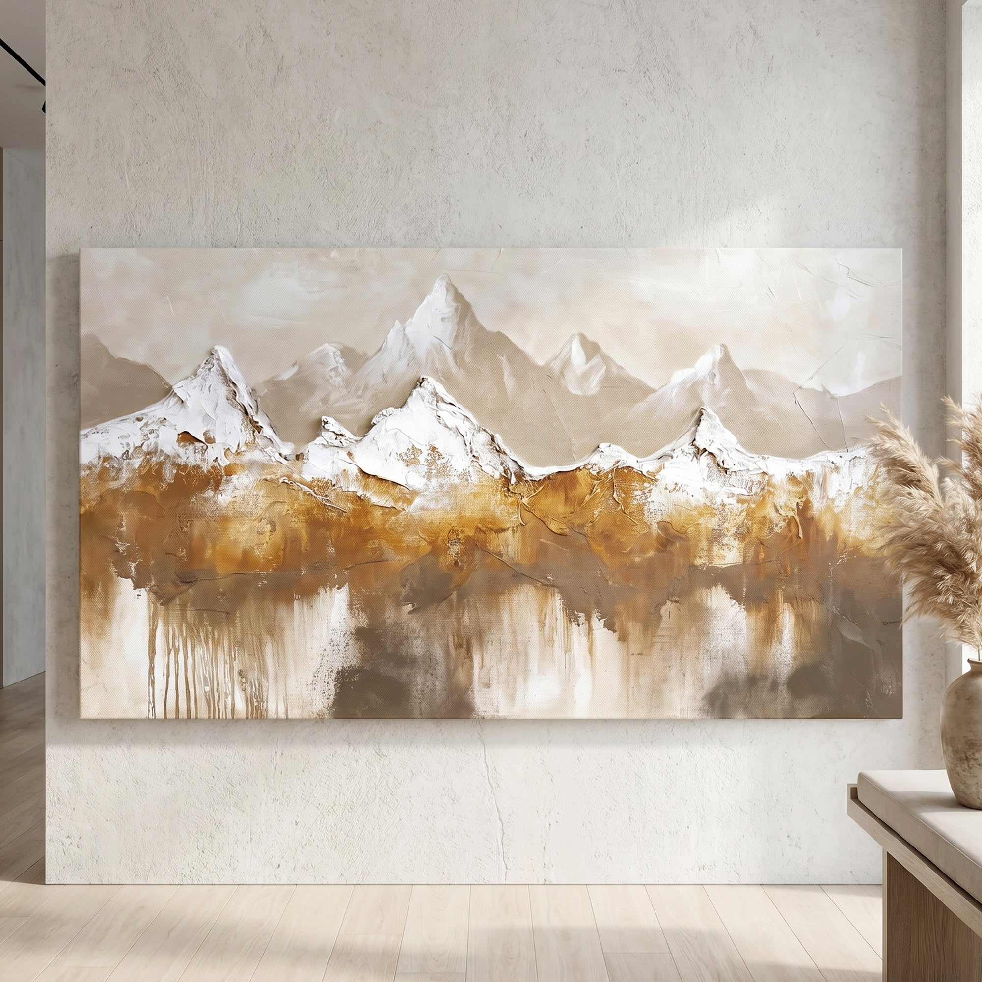 A large rectangular canvas painting of a mountain landscape hangs on a textured light gray wall in a styled interior. The artwork shows snow-capped peaks above golden-brown and beige reflective water, with a vase and dried grasses at the right edge.