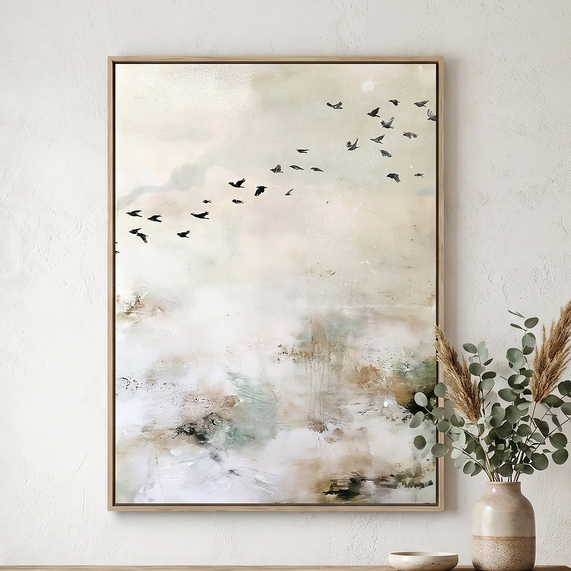 Framed abstract wall art hanging on a textured white wall, featuring a soft cream and beige composition with small black birds flying across the upper right and a light wood frame. A beige vase with dried grass and greenery sits below the frame.
