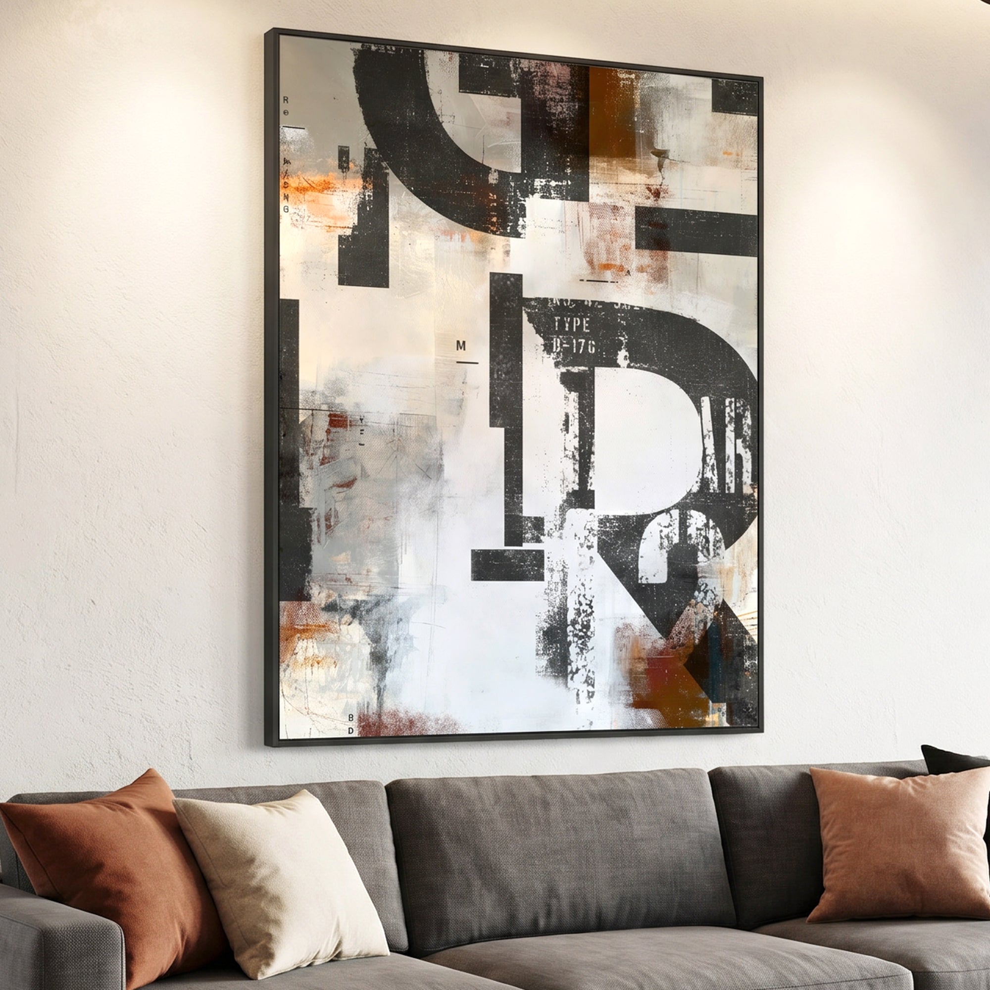 Large framed abstract wall art with bold black, white, gray, and rust-orange letterforms, displayed above a gray sofa with brown and cream pillows in a real interior setting.