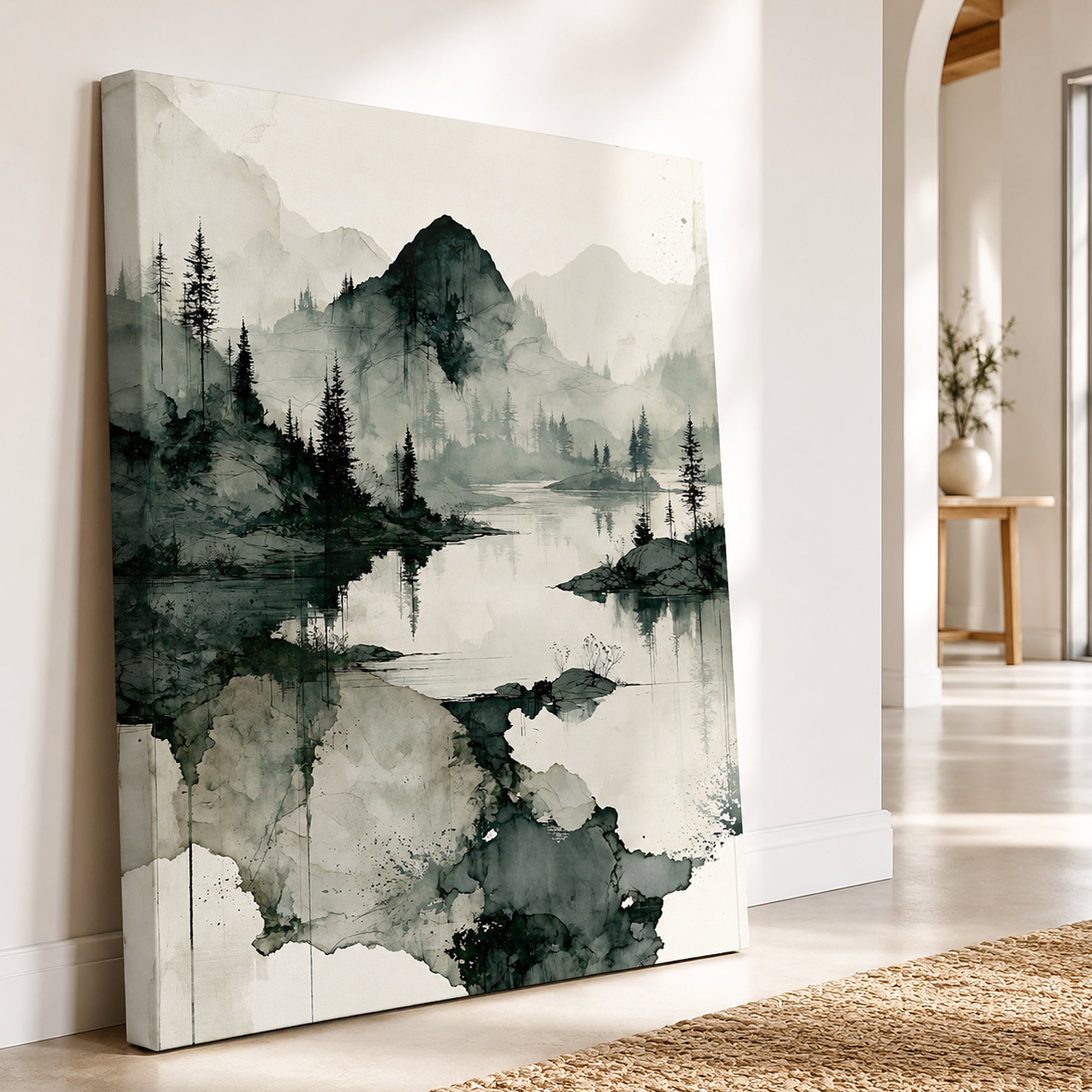 A vertical canvas wall art print featuring a serene black and white ink-style landscape with mountains, trees, and reflective water, leaning against a white wall in a bright modern room.