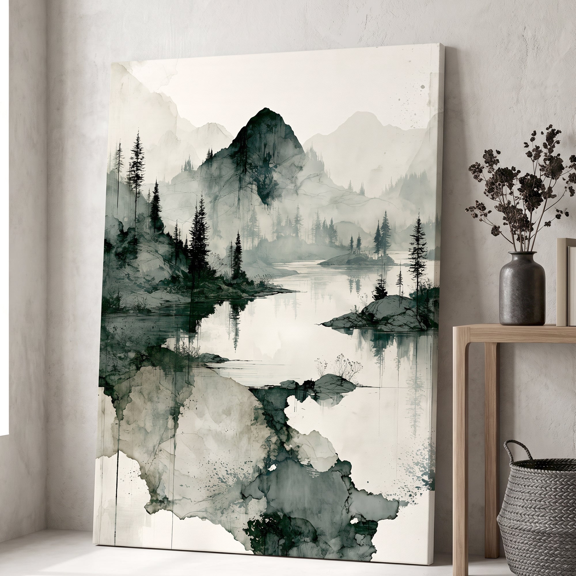 A vertical canvas wall art print featuring a serene landscape with misty mountains, pine trees, and a reflective lake in dark green, gray, and off-white tones, styled leaning against a textured wall next to a wooden side table with a vase of dried stems.